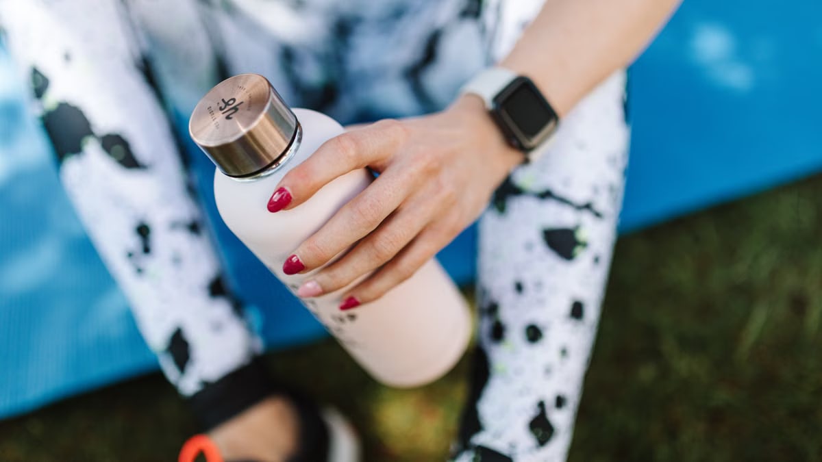 Crop unrecognizable female wearing sports leggings smart watch and holding bottle with water while relaxing after training on mat outside